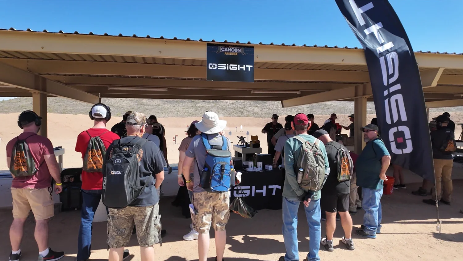 Outdoor shooting range event with a group of people gathered under a canopy, wearing backpacks and ear protection, with a banner overhead.